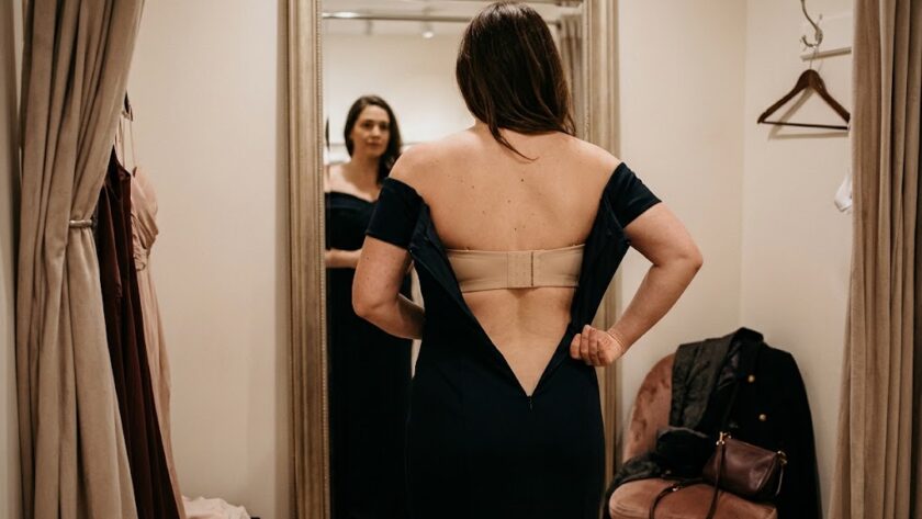 Woman wearing an off-the-shoulder evening dress checking strapless bra support in front of a boutique mirror